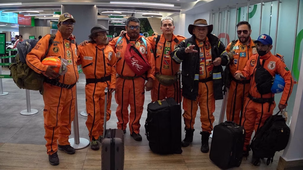 Grupo de siete rescatistas mexicanos conocidos como "Los Topos" posando en un aeropuerto. Visten sus característicos overoles naranjas con arneses negros y parches internacionales, junto a sus maletas y cascos de seguridad.