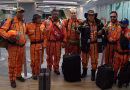 Grupo de siete rescatistas mexicanos conocidos como "Los Topos" posando en un aeropuerto. Visten sus característicos overoles naranjas con arneses negros y parches internacionales, junto a sus maletas y cascos de seguridad.