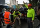 El rescatista Jorge Beens, vestido con camisa amarilla de manga larga y arnés de rescate, acaricia al perro de búsqueda Tsunami mientras este descansa en la plataforma de una camioneta pickup negra en La Guaira. A la derecha, otro rescatista con casco naranja sostiene en alto una bolsa de suero intravenoso conectada al perro. En el fondo se aprecian edificios multifamiliares y estructuras colapsadas por el terremoto.