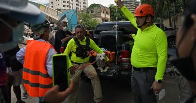 El rescatista Jorge Beens, vestido con camisa amarilla de manga larga y arnés de rescate, acaricia al perro de búsqueda Tsunami mientras este descansa en la plataforma de una camioneta pickup negra en La Guaira. A la derecha, otro rescatista con casco naranja sostiene en alto una bolsa de suero intravenoso conectada al perro. En el fondo se aprecian edificios multifamiliares y estructuras colapsadas por el terremoto.