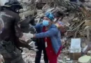 Un médico veterinario con guantes, mascarilla quirúrgica y gorro médico recibe entre sus manos a la perrita negra Luz Clarita, quien le es entregada por un funcionario de rescate uniformado. En el fondo se aprecia la zona cero cubierta por montañas de escombros, hierros y concreto colapsado.