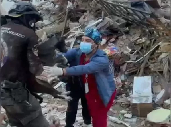 Un médico veterinario con guantes, mascarilla quirúrgica y gorro médico recibe entre sus manos a la perrita negra Luz Clarita, quien le es entregada por un funcionario de rescate uniformado. En el fondo se aprecia la zona cero cubierta por montañas de escombros, hierros y concreto colapsado.